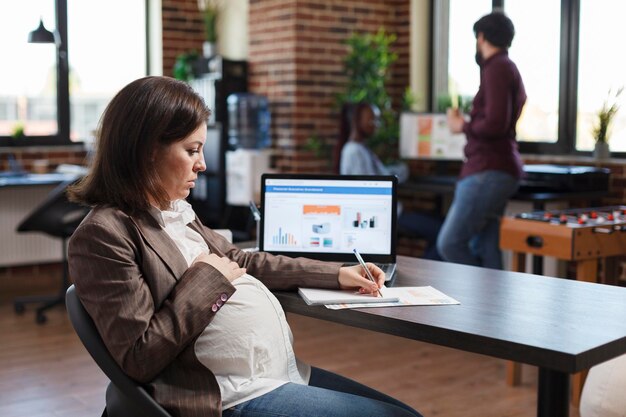 Mujer embarazada que analiza el crecimiento económico de la empresa y revisa el gráfico financiero y de gestión. Empleado de la agencia financiera que revisa el desarrollo del proyecto mientras mira el gráfico en el monitor.