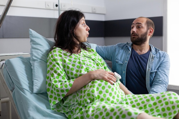 Mujer embarazada que se pone de parto en la sala del hospital, esposo de pie junto a ella reconfortante. Paciente con embarazo cogido de la mano en el vientre haciendo ejercicios de respiración mientras tiene contracciones dolorosas