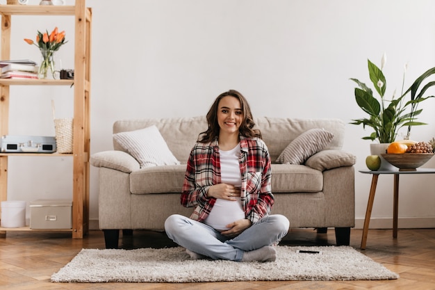 Foto gratuita mujer embarazada rizada con hermoso maquillaje vestida con camisa a cuadros y jeans toca suavemente el vientre y sonríe sinceramente mientras está sentada en una alfombra beige suave cerca del sofá en la acogedora habitación