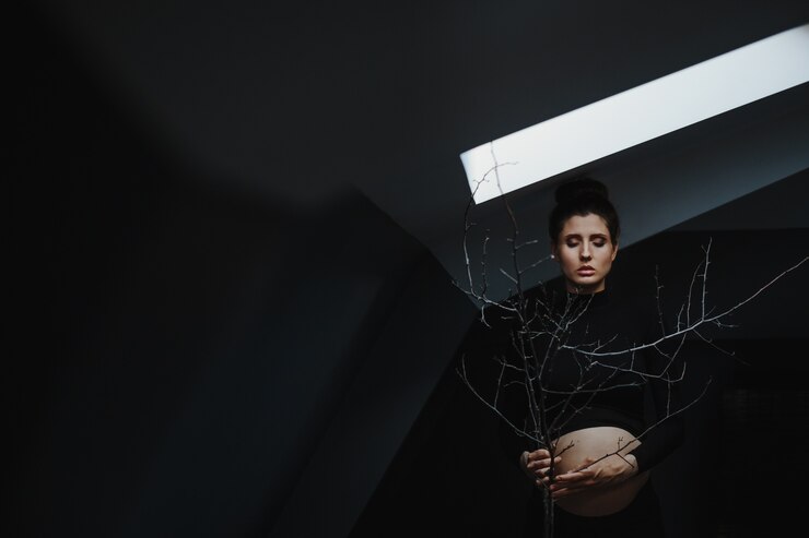 Foto gratuito la mujer embarazada en ropa negra se coloca debajo de la pared gris y sostiene una ramificación de árbol