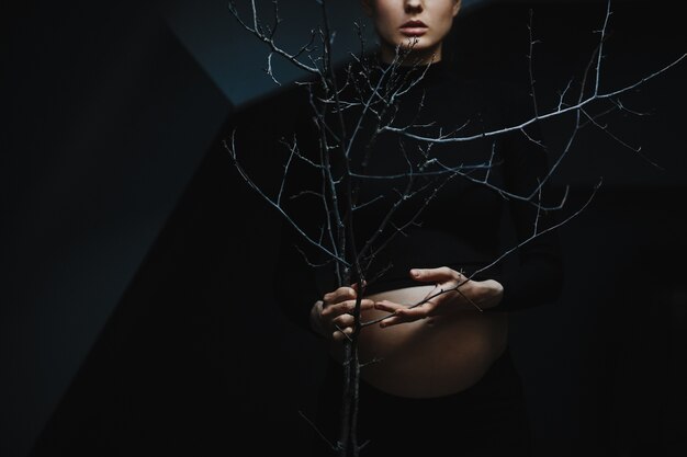 La mujer embarazada en ropa negra se coloca debajo de la pared gris y sostiene una ramificación de árbol