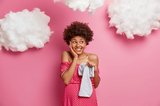 Mujer embarazada satisfecha con cabello afro mantiene la mano en las mejillas sostiene body de bebé sonríe agradablemente usa vestido tiene poses de abdomen grande