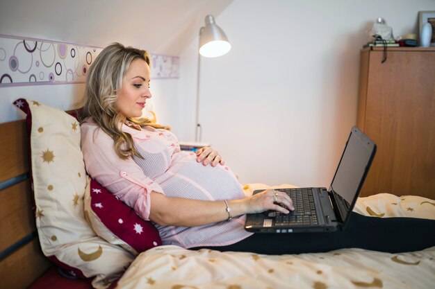 Mujer embarazada sentada en la cama usando la computadora portátil