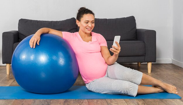 Mujer embarazada sonriente en casa entrenando con pelota y usando teléfono móvil