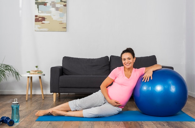 Mujer embarazada sonriente en casa con pelota y colchoneta de ejercicios