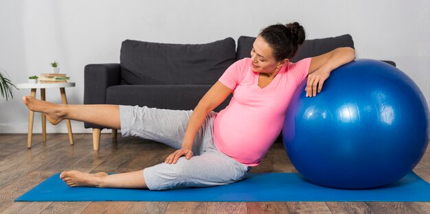 Mujer embarazada sonriente en casa con pelota de ejercicios y colchoneta