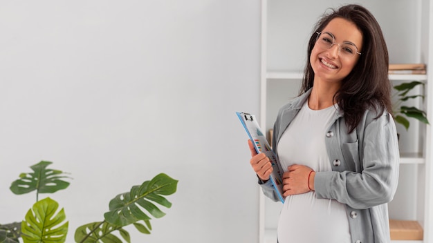 Mujer embarazada sonriente en casa trabajando mientras sostiene el portapapeles