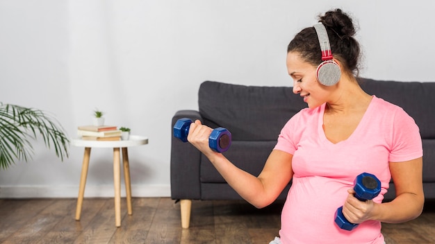 Foto gratuita mujer embarazada sonriente escuchando música en auriculares mientras hace ejercicio con pesas