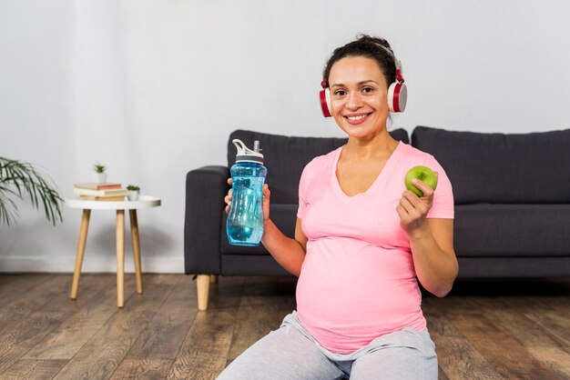 Mujer embarazada sonriente escuchando música en auriculares mientras sostiene la botella de agua y manzana