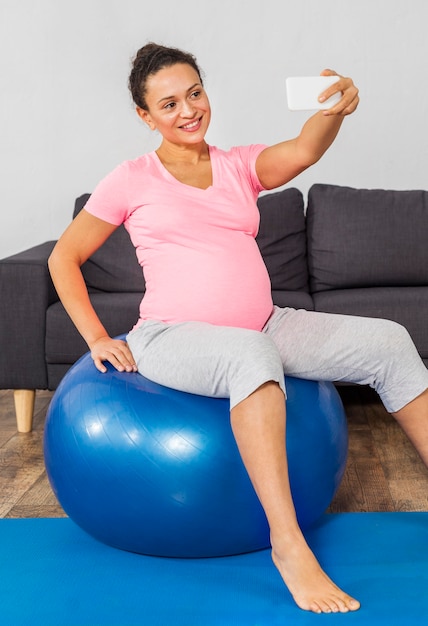 Mujer embarazada sonriente tomando selfie en casa mientras entrena con pelota