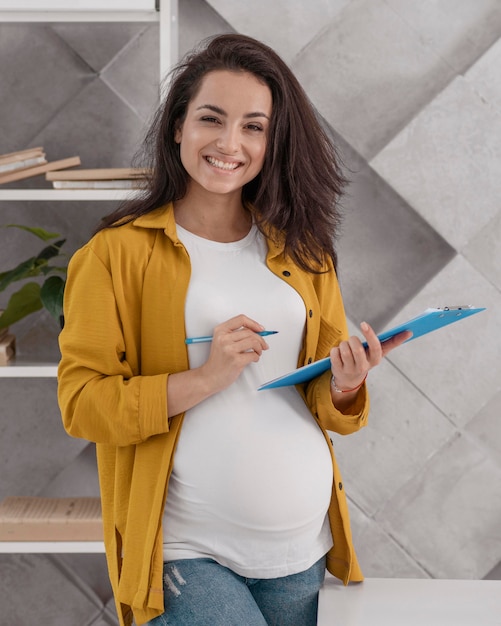 Mujer embarazada sonriente trabajando desde casa con portapapeles y lápiz