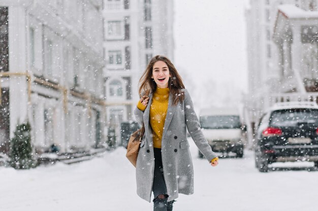 Mujer emocionada en abrigo gris y jeans rotos caminando por la calle en un día de nieve. Mujer caucásica de moda pasar tiempo al aire libre en invierno, explorando la ciudad.