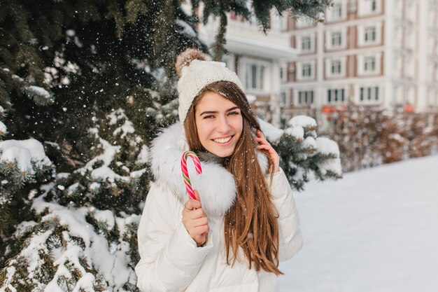 Mujer emocionada con cabello castaño lacio divirtiéndose en un día de nieve y disfrutando de la sesión de fotos. Retrato al aire libre de la impresionante dama blanca en ropa de moda posando con dulces navideños.