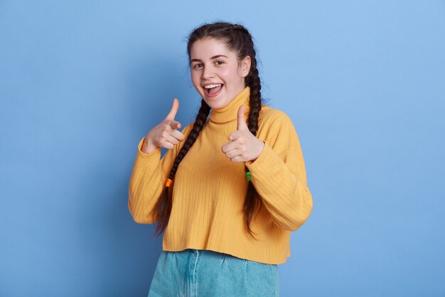 Mujer emocionada con coletas apuntando con los dedos índices, mirando con sonrisa encantadora, tiene emociones positivas, de pie aislado.