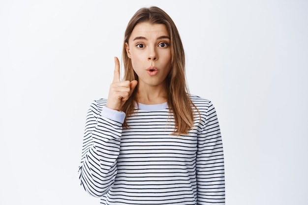 Mujer emocionada comparte pensamientos, levanta el dedo y tiene un plan, agrega sugerencia o solución de maquillaje, de pie sobre una pared blanca