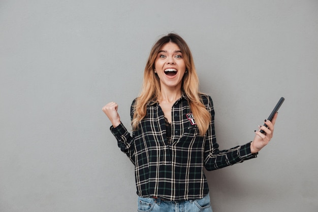 Mujer emocionada feliz que sostiene el teléfono móvil y que celebra victoria