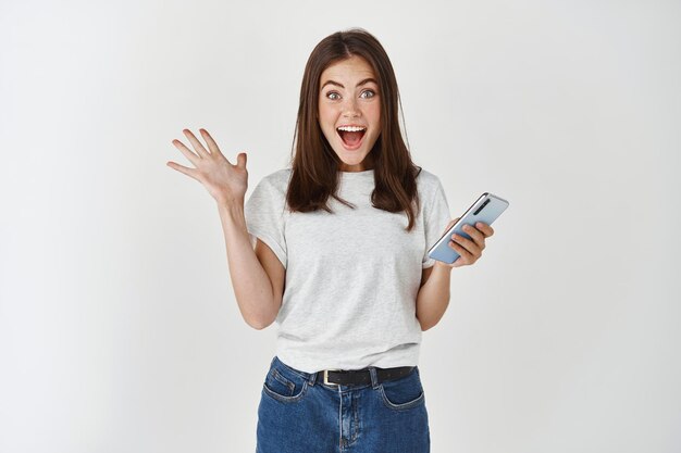 Mujer emocionada ganando el premio en el teléfono, regocijándose y mirando al frente feliz, grito de alegría sobre la pared blanca.