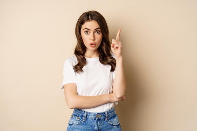 Mujer emocionada levantando el dedo, lanzando una idea, tiene sugerencia, usando camiseta blanca y jeans sobre fondo beige.