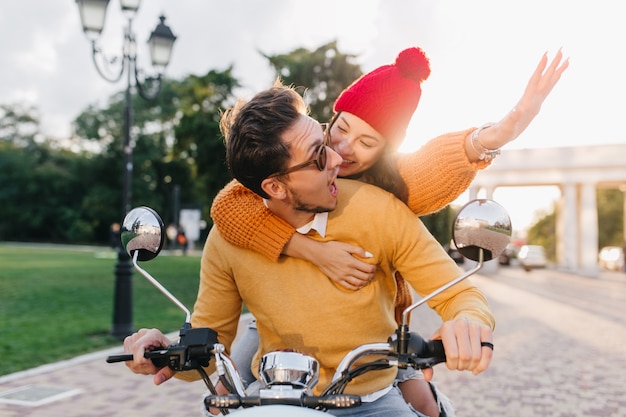 Mujer emocionada lleva sombrero colorido abrazando al hombre mientras conduce scooter