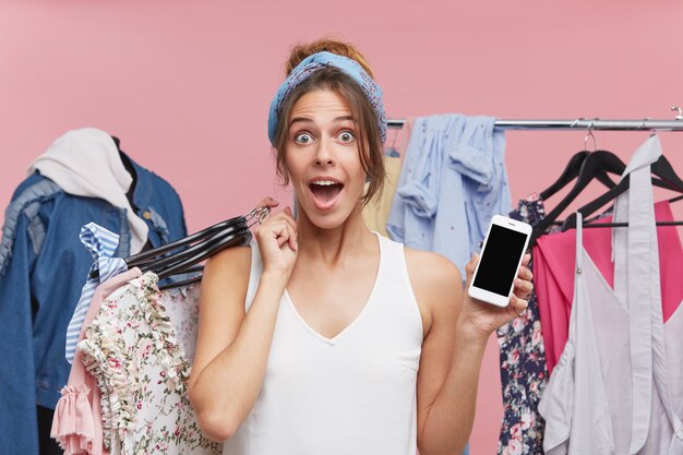 Mujer emocionada mirando con gran sorpresa, sosteniendo perchas con ropa mientras está de pie en el guardarropa, demostrando el teléfono móvil con pantalla en blanco. Gente, compras, concepto de tecnología