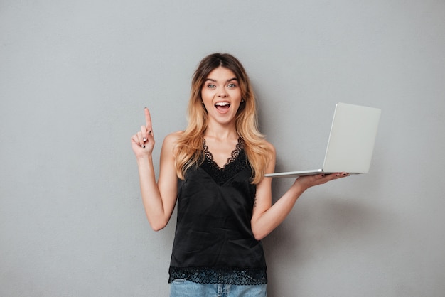 Mujer emocionada sosteniendo portátil y apuntando el dedo hacia arriba en copyspace