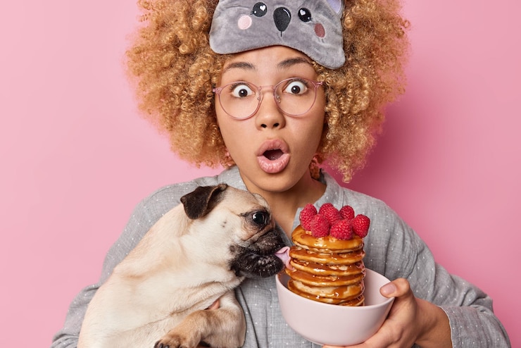 Foto gratuito mujer emocional asustada con cabello rizado mira fijamente los ojos saltones tiene la boca abierta por el shock sostiene un perro pug que lame panqueques hechos para el desayuno vestido con ropa de dormir aislado en la pared rosa