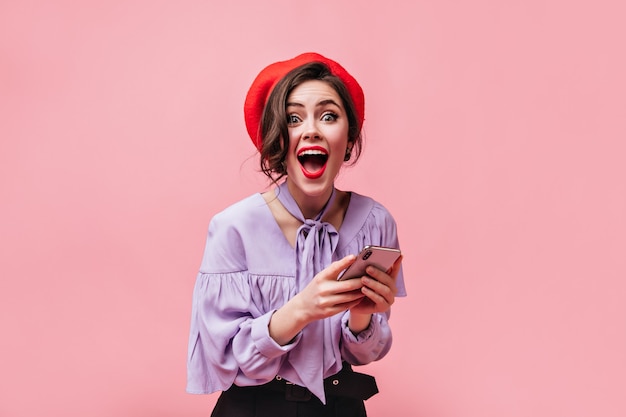 Mujer emocional en boina roja y camisa elegante en alegre sorpresa mira a cámara y sostiene teléfono inteligente blanco sobre fondo rosa.