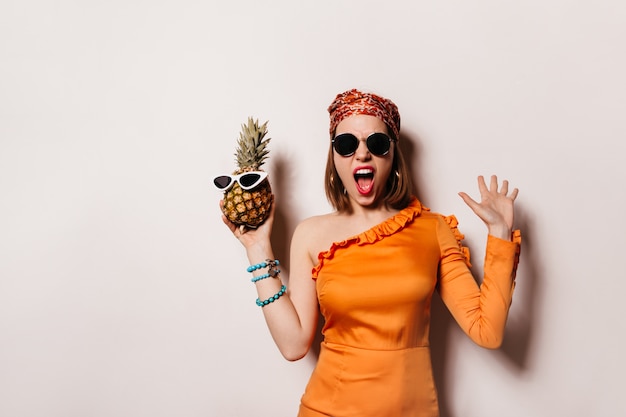 Mujer emocional con elegante diadema y vestido naranja pretenciosamente grita y sostiene piña en gafas de sol.