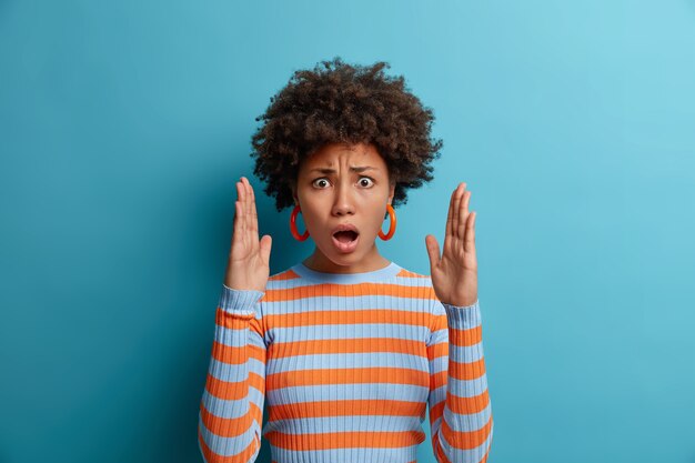 Mujer emocional estupefacta impresionada por el gran tamaño, levanta las palmas y muestra algo enorme, abre la boca y explica, da forma a un objeto invisible, extiende los brazos, vestida con un jersey de rayas, aislado en azul