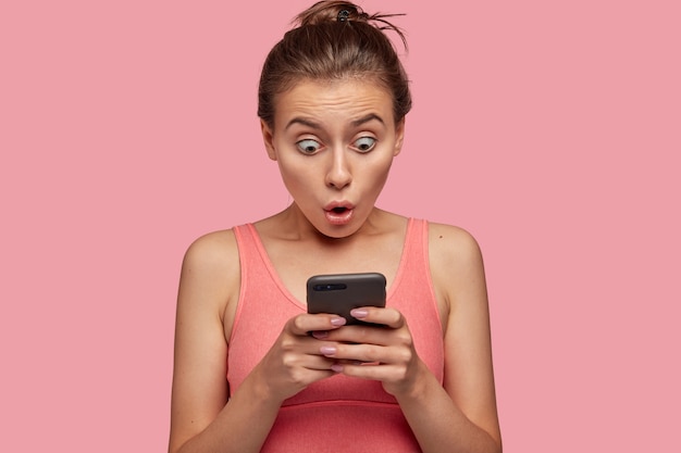 Mujer emocional estupefacta mira fijamente con ojos saltones al teléfono inteligente, se siente sorprendida mientras lee noticias en la página web, está conectada a internet inalámbrico, tiene un descanso después de entrenar con el entrenador en el gimnasio, usa una camiseta rosa