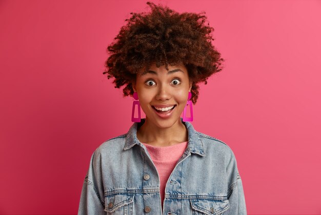 Mujer emocional feliz sorprendida con cabello afro se ve con una sonrisa, no puede creer que los sueños se hagan realidad, recibe un regalo increíble de alguien, vestida con ropa de mezclilla de moda, aislada en una pared rosa