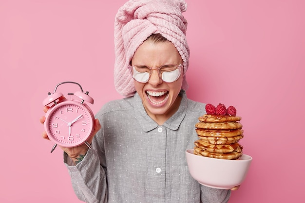 Foto gratuita la mujer emocional grita en voz alta mantiene la boca abierta se despierta muy temprano sostiene el despertador y los panqueques apetitosos se viste con pijama aplica parches de belleza debajo de los ojos para eliminar la hinchazón después de dormir