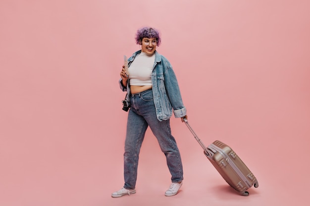 Mujer emocional en jeans y zapatillas blancas tiene boletos y maleta gris. Señora con gafas y top ligero riendo en rosa.