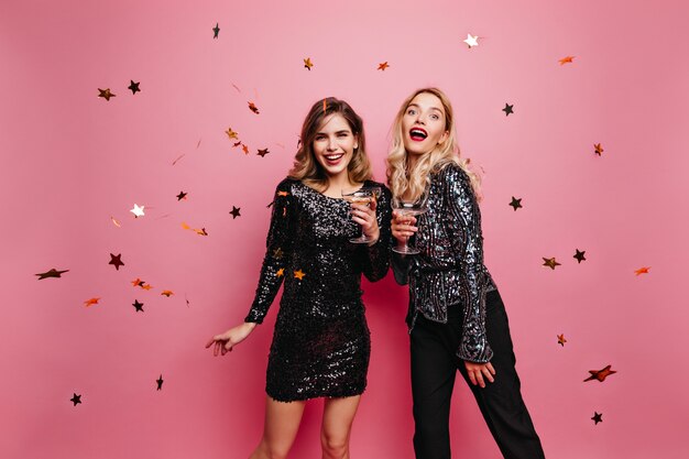 Mujer emocional en pantalón negro y chaqueta brillante bebiendo vino. Encantadoras amigas disfrutando de la fiesta de cumpleaños.