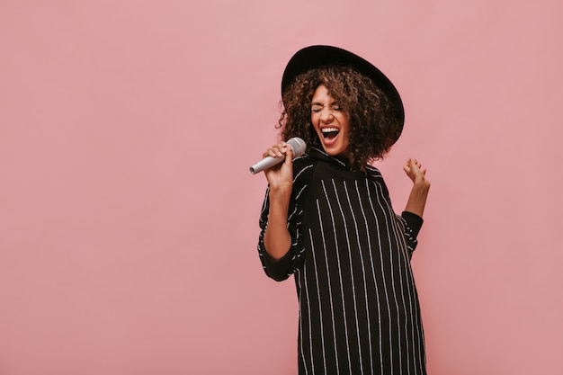 Mujer emocional con peinado morena rizado en elegante sombrero y vestido negro a rayas sosteniendo el micrófono y cantando en la pared rosa.