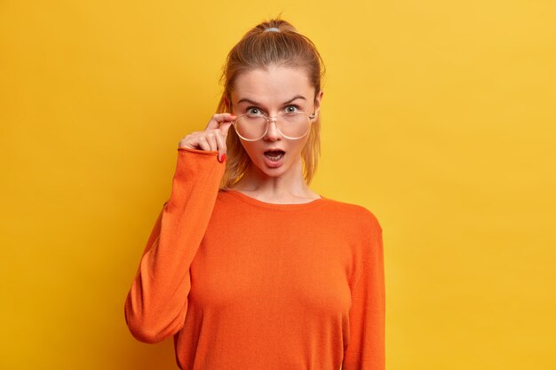 Mujer emocional sorprendida con la boca ampliamente abierta mira a través de gafas ópticas, vestida con un jersey naranja casual, escucha noticias asombrosas, posa