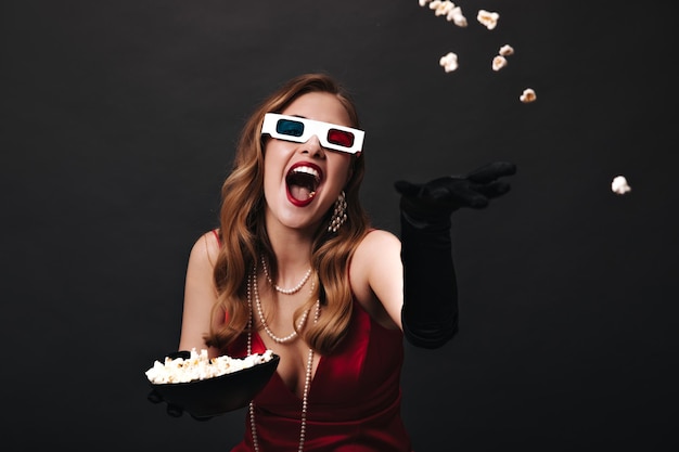 Mujer emocional en traje festivo y gafas 3D lanzando palomitas de maíz Encantadora dama feliz en la parte superior de seda roja viendo la película sobre fondo negro