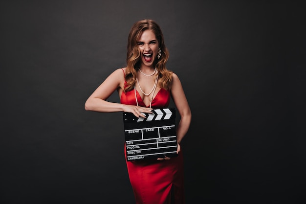 Foto gratuita mujer emocional en vestido rojo sonriendo y sosteniendo claqueta mujer feliz rubia en traje brillante satinado riendo sobre fondo negro