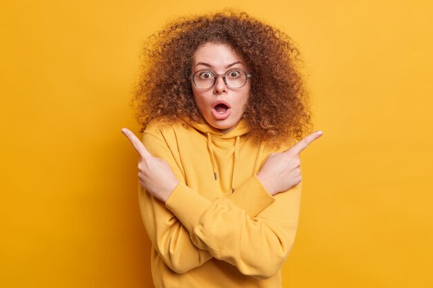 Una mujer emotiva aterrorizada conmocionada con el pelo rizado apunta hacia los lados en variantes indica que la derecha y la izquierda mantienen la boca bien abierta vestida con una sudadera con capucha no pueden elegir entre dos elementos aislados en la pared amarilla