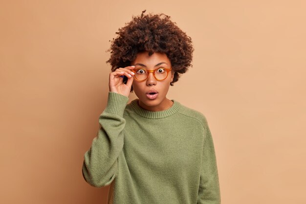 La mujer emotiva mira fijamente al frente tiene los ojos abiertos contiene el aliento de la maravilla mira escrupulosamente al frente usa un suéter casual aislado sobre una pared beige