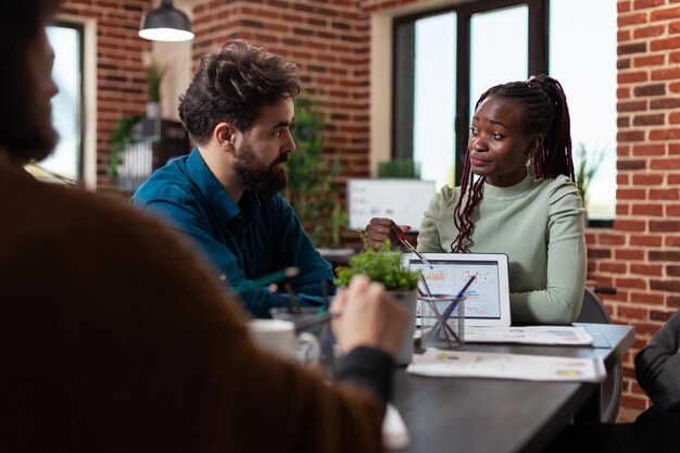 Mujer empresaria afroamericana que muestra estadísticas de marketing a empresarios que explican el volumen de negocios de la empresa durante una reunión de negocios en la oficina de inicio. Diverso equipo trabajando en proyecto financiero