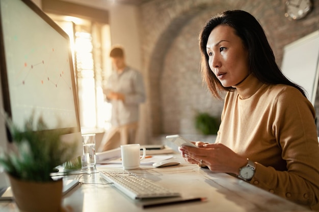 Mujer empresaria asiática leyendo gráficos en el monitor de la computadora y enviando mensajes de texto en un teléfono inteligente en la oficina