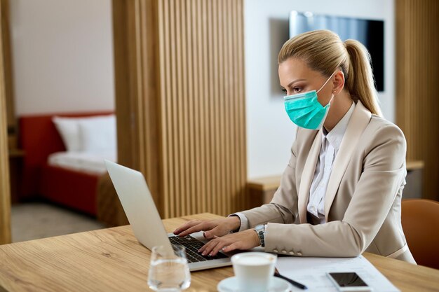 Mujer empresaria con mascarilla protectora escribiendo un correo electrónico en una computadora en la habitación del hotel