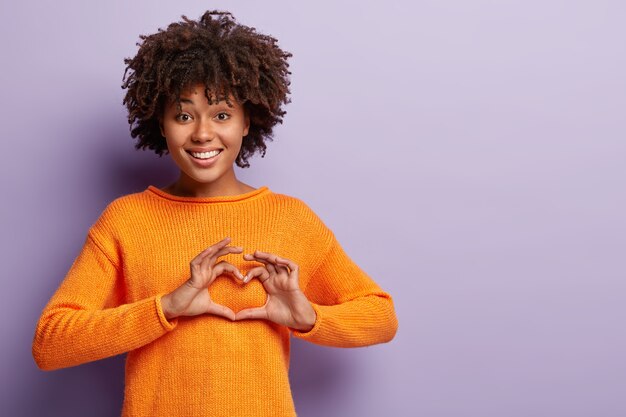 Mujer encantadora afectuosa tiene el corazón lleno de amor, muestra el signo de San Valentín, vestida con un jersey naranja casual