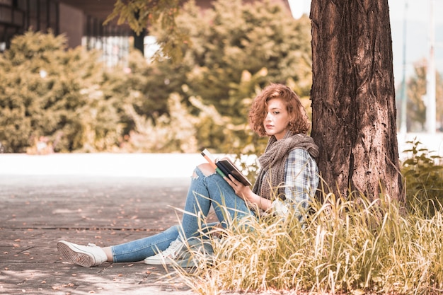 Foto gratuita mujer encantadora apoyándose en el árbol y sosteniendo el libro en el parque