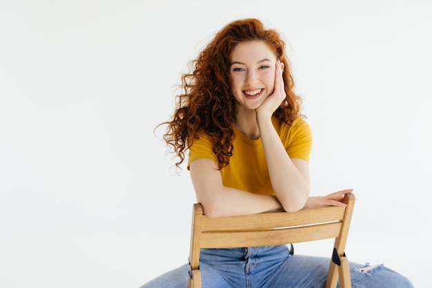 Foto gratuita mujer encantadora confiada en jeans azules posando en una silla foto de estudio de una dama caucásica de jengibre sentada cerca de una pared blanca