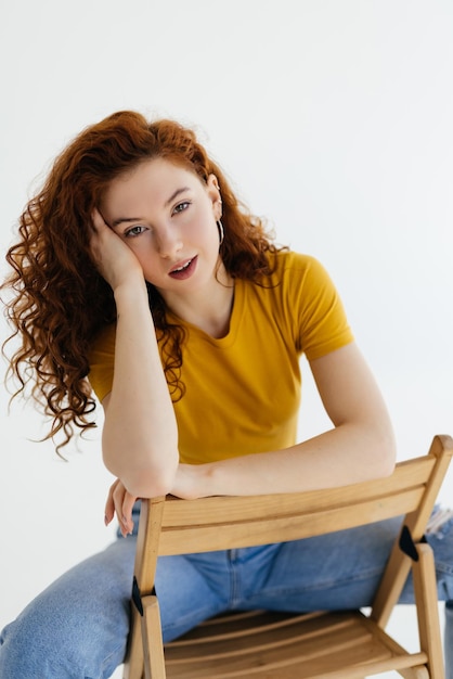 Mujer encantadora confiada en jeans azules posando en una silla Foto de estudio de una dama caucásica de jengibre sentada cerca de una pared blanca