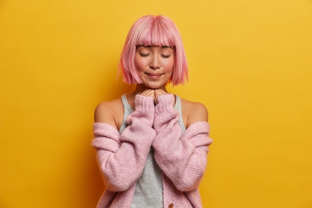 Foto gratuita una mujer encantadora conmovida tiene un peinado bob rosa y flecos, se para en el interior, cierra los ojos y mantiene las manos debajo del mentón