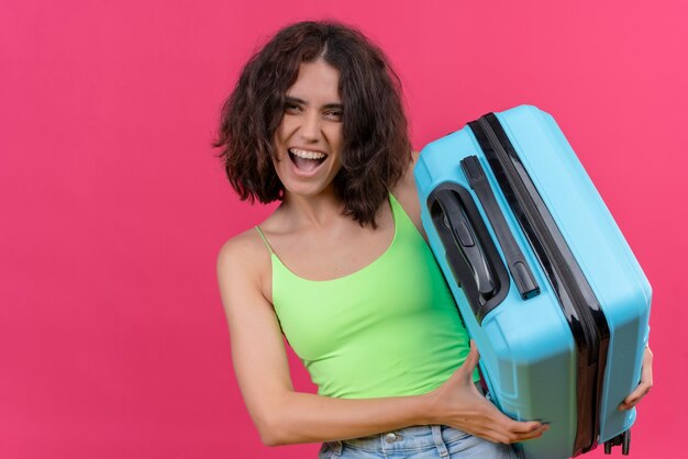 Una mujer encantadora feliz con el pelo corto vistiendo verde crop top mostrando su maleta azul