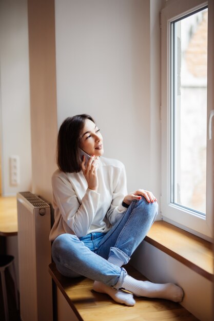 Mujer encantadora en la habitación sentada cerca de la ventana en ropa casual suéter blanco